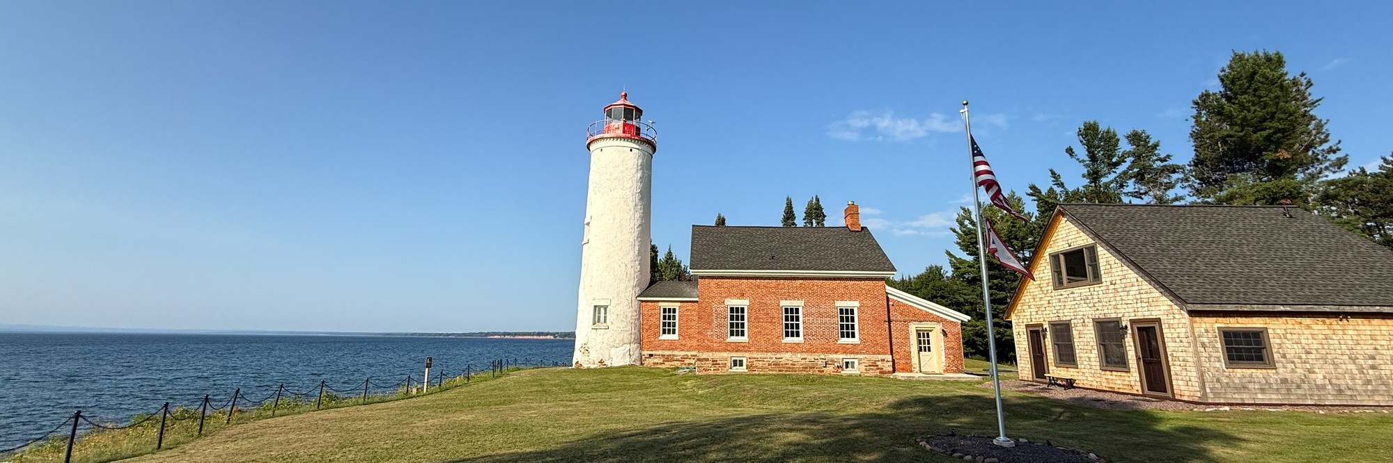 The Portage River light station in summer, white tower and red brick keeper's house on a mown green point above Lake Superior, flag flying beside a shingled outbuilding