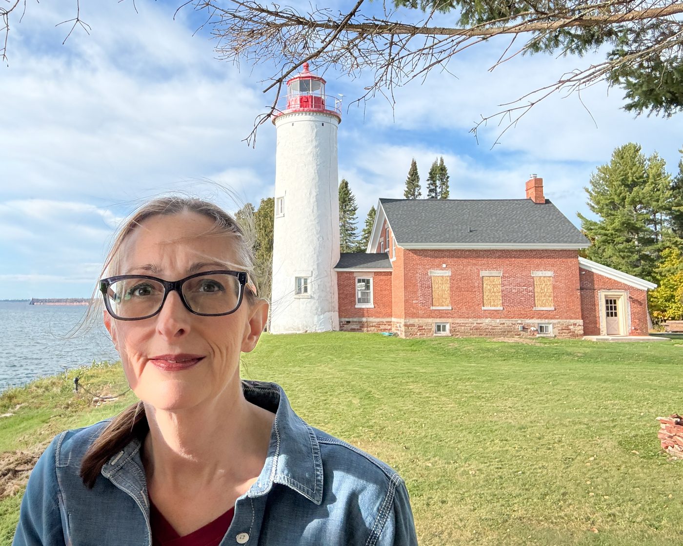 Susan Harbourt outside the Portage River light station, the white tower and red brick keeper's house behind her on a green point above Lake Superior